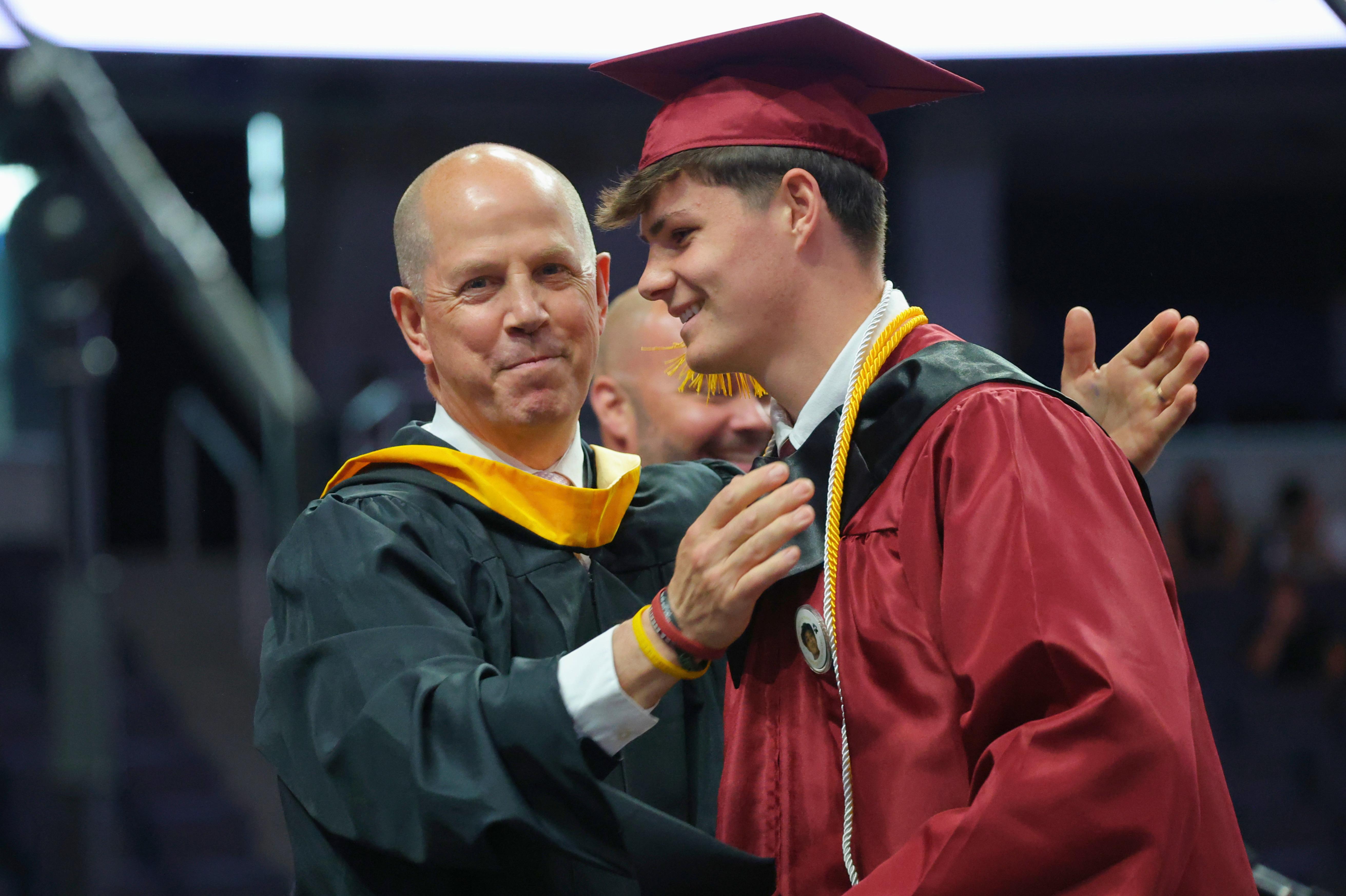 A man in academic regalia pats a boy wearing a red cap and gown on the back